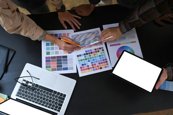 Hände, die an einem Tisch mit Farbmuster, Dokumenten und einem Tablet arbeiten.