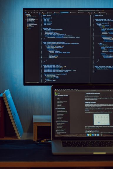 Ein Arbeitsplatz mit einem Laptop und einem großen Monitor, beide zeigen Programmiercode.