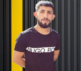 Junger Mann mit Bart in schwarzem T-Shirt vor einem gestreiften Hintergrund.