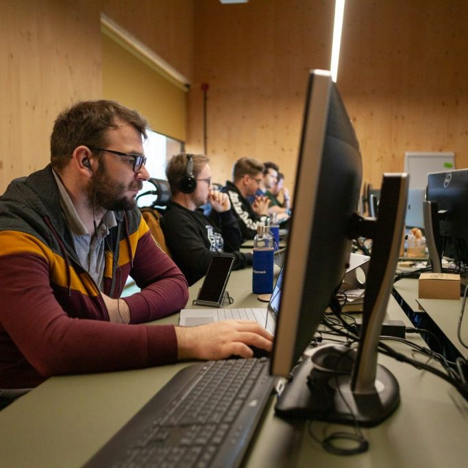 Person am Computer, umgeben von weiteren Personen in einem Büro mit Holzverkleidung.