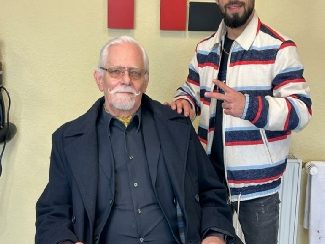 Ein älterer Mann im Frisörstuhl, umarmt von einem jüngeren Mann, beide lächeln.