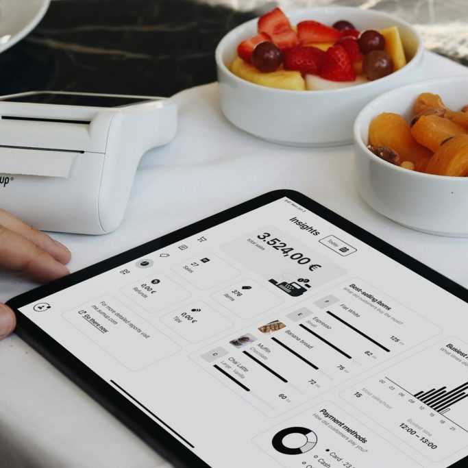 Tablet mit Datenanzeige neben Schalen mit Obst und Joghurt auf einem Tisch.